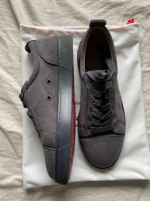 Gråa sneakers i mocka med röd sula - Snyggare sneakers i grå mocka med mörkgrå sula och cool röd undersida. Skorna har klassisk låg profil, svarta skosnören och metallöljetter på sidan. Perfekta för dig som gillar stilrena och moderna detaljer.