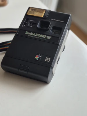 Kodak EK160-EF instantkamera - Retro svart instantkamera från Kodak, modell EK160-EF. Kameran har inbyggd elektronisk blixt och en regnbågsfärgad handledsrem. Tillverkad i plast med klassisk 80-talsdesign och tydliga logotyper på framsidan.
