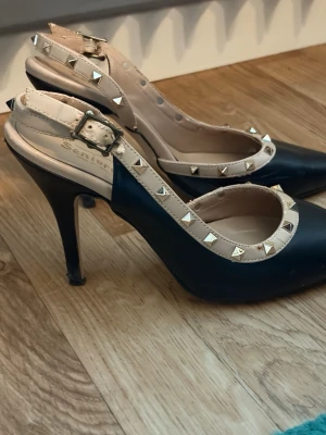Svarta pumps med nitar och beige kant - Snygga svarta pumps från Santos med spetsig tå och höga klackar. Skorna har beige kant med coola guldfärgade nitar och justerbar rem runt ankeln. Perfekta för dig som gillar en edgy och trendig stil.