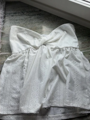 Vit axelbandslös broderad topp - Söt vit axelbandslös topp med knutdetalj framtill och broderade hålmönster. Toppen har en luftig passform och är perfekt för varma dagar. Tillverkad i bomull för en skön känsla och stilren look.