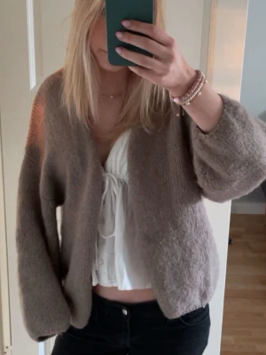 Brun Lindex kofta  - Mysig brun kofta med fluffig känsla och ballongärmar. Koftan har en avslappnad passform och är öppen framtill utan knappar. Perfekt att slänga över en topp för en chill look.