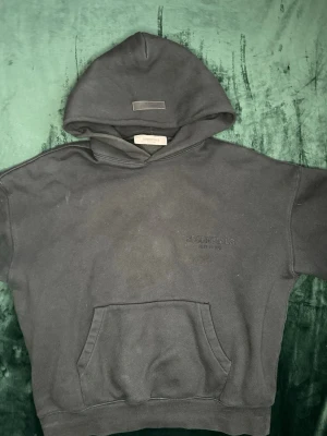 Svart track suit hoodie från Fear of God Essentials - Säljer en brun hoodie från Fear of God Essentials med broderad logga på bröstet. Hoodien har en stor känguruficka framtill, huva och avslappnad passform. Perfekt för dig som gillar streetwear och vill ha en stilren look. Om du skulle vilja köpa bara hoodien kostar de 1000 och byxor 700 men både för 1500 eller mindre. Pris kan diskuteras. MVH stentorparchive⚓️