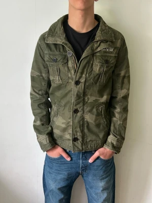 Camouflage jacka från Hollister - Säljer en grön camouflage jacka från Hollister med klassisk krage och knappar framtill. Jackan har två bröstfickor med knappstängning och två sidofickor. Perfekt för dig som vill ha en cool och avslappnad stil.
