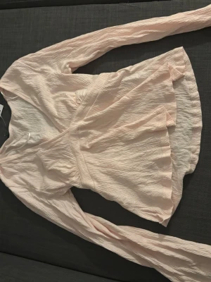 Ljusrosa blus   - Supersöt ljusrosa omlottblus med lång ärm och v-ringning. Blusen har en lätt skrynklig struktur och markerad midja som ger en snygg siluett. Perfekt att styla med jeans eller kjol för en trendig look. Knyts bak för att framhäva midjan. Aldrig använd och har lappen kvar.