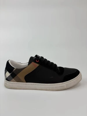 Svarta sneakers från Burberry - Stilrena svarta sneakers från Burberry med klassiskt rutigt mönster i beige, svart och vitt på sidan. Skorna har snörning och vit sula. Perfekta för dig som vill ha en snygg och tidlös look med en touch av lyx.