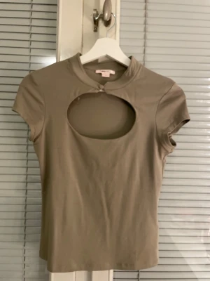 Grön topp med cut-out och knapp - Trendig grön topp med korta ärmar och en stor cut-out detalj framtill. Toppen har en liten ståkrage med knappstängning vid halsen. Perfekt för dig som vill sticka ut med en unik detalj. Mjuk och stretchig känsla.