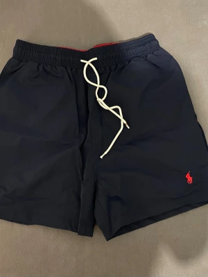 Polo Ralph Lauren Badshorts   - Snygga marinblå badbyxor från Polo Ralph Lauren med en bakficka med lock. Perfekta för stranden eller sommaren. Storlek M men passar mer till S 