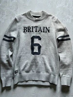 Grå stickad morris tröja - Grå stickad tröja från morris med svart text 'BRITAIN' och siffran 6 framtill. Två svarta ränder på ärmarna och ribbstickade muddar vid hals, ärmslut och nederkant. Perfekt för en sportig och avslappnad stil.