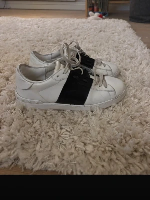 Vita sneakers med svart band Valentino - Snygga vita sneakers från Valentino med ett brett svart band över mitten och vita snören. Skorna har en låg profil och detaljerade nitar på sulan. Tillverkade i läder med klassisk rund tå och stilren design.