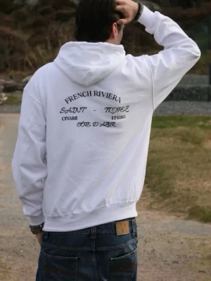 Vit hoodie French Riviera print - Vit hoodie med svart text på ryggen där det står 'French Riviera', 'Saint Tropez', 'Côte d’Azur' och 'Studio'. Klassisk passform med huva och ribbade muddar. Perfekt för en chill och stilren look. Sitter M men sitter även snyggt på s om man har långa ärmar