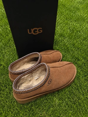 Bruna UGG tofflor i mocka - Säljer ett par bruna UGG tofflor i mjuk mocka med fluffigt foder och snyggt mönstrad kant i vitt och gult. Perfekta för att hålla fötterna varma hemma. Klassisk UGG-logga på sidan och slitstark sula.
