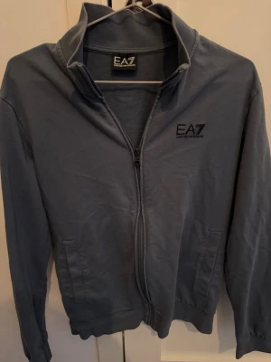 Grå zip-hoodie från EA7 Emporio Armani - Snygg grå hoodie med dragkedja från EA7 Emporio Armani. Tröjan har två fickor framtill, ribbade muddar och hög krage. Perfekt för en avslappnad och stilren look. Materialet känns mjukt och bekvämt mot huden.