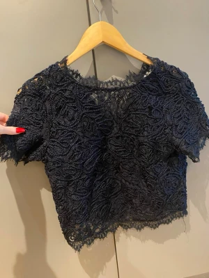 Mörkblå spetsblus med kort ärm - Snygg mörkblå blus i spets med korta ärmar och rund halsringning. Blusen har ett vackert broderat mönster och är lätt transparent. Perfekt att styla med höga jeans eller kjol för en trendig look.
