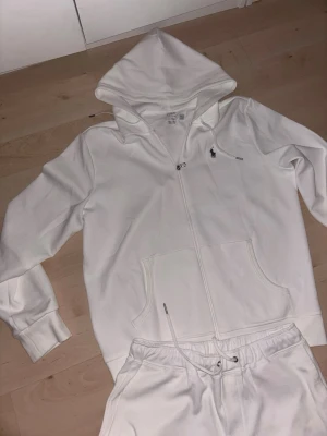 Vit tracksuit från Ralph Lauren - Snyggt vitt loungewear-set från Ralph Lauren med hoodie och matchande byxor. Hoodien har dragkedja, fickor och den klassiska loggan på bröstet. Byxorna har dragsko i midjan och logga på benet. Perfekt för chill dagar hemma.