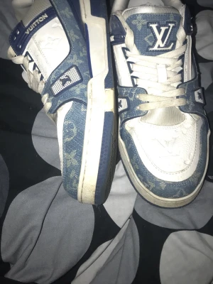 Louis Vuitton blåvita sneakers - Säljer ett par Louis Vuitton sneakers i blått och vitt med klassiskt monogrammönster. Skorna har vita snören, robust sula och LV-logga på plösen. Materialet är en mix av denim och läder, vilket ger en unik och lyxig känsla. Perfekta för dig som vill sticka ut med exklusiva detaljer.
