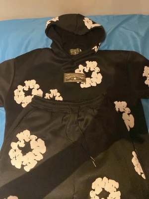 Svart loungewear-set från Denim Tears - Snyggt svart loungewear-set från Denim Tears med vita blomliknande tryck över hela både hoodie och byxor. Hoodien har huva och en patch framtill. Perfekt för chill dagar hemma eller när du vill vara bekväm men ändå se cool ut.