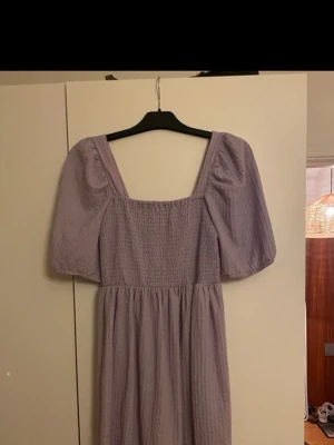 Lavendel lila kortklänning med puffärm - Supersöt lavendel lila kortklänning med puffärmar och smockad överdel. Klänningen har fyrkantig ringning och är tillverkad i ett lätt, strukturerat tyg som ger en luftig känsla. Perfekt för varma dagar och trendiga outfits. Endast använd en gång och är väldigt fin utan defekter och skavanker.