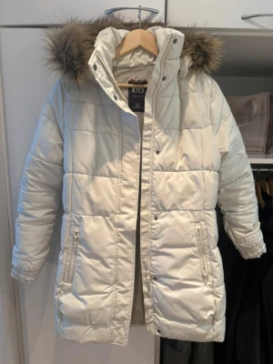 Vit dunjacka med pälskant på huvan - Säljer en vit dunjacka från Bik Bok med avtagbar huva och pälskant. Jackan har två dragkedjeförsedda fickor fram, quiltad design och är perfekt för kalla vinterdagar. Stängs med dragkedja och knappar. Snygg och varm modell som håller dig bekväm.