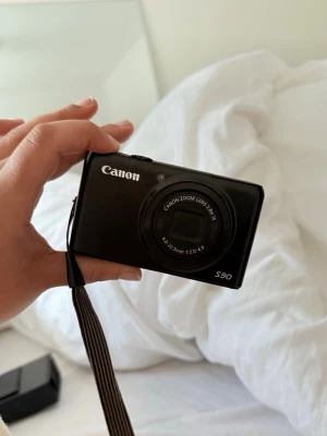 Canon PowerShot S90 digitalkamera - Säljer en svart Canon PowerShot S90 kompakt digitalkamera med inbyggd zoomlins (6.0-22.5mm f/2.0-4.9). Kameran har en smidig rektangulär form, tydliga reglage på ovansidan och en praktisk handledsrem. Perfekt för dig som vill ha en enkel och stilren kamera att ta med överallt.