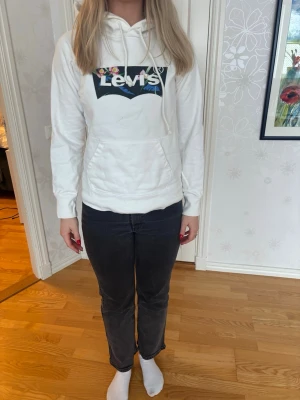 Vit hoodie från Levi's med logga - Säljer en vit hoodie från Levi's med stor färgglad logga på bröstet. Hoodien har huva med dragsko och en klassisk magficka framtill. Tillverkad i mjukt material och har ribbade muddar vid ärmar och nederkant. Perfekt för en avslappnad och trendig look.