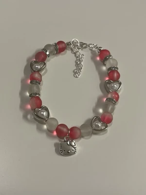 Hello Kitty armband med pärlor -  HANDGJORT armband med rosa och vita pärlor, silverfärgade hjärtan och en Hello Kitty-berlock. Armbandet har justerbar kedja.