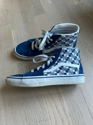 Blå Vans Sk8-Hi sneakers strl 36 - Säljer ett par blå Vans Sk8-Hi sneakers i storlek 36 med vit snörning och checkerboard-mönster med Vans-logga på sidan. Skorna har mockadetaljer och klassisk vit sula med blå rand. Höga och riktigt snygga för dig som gillar streetstyle.