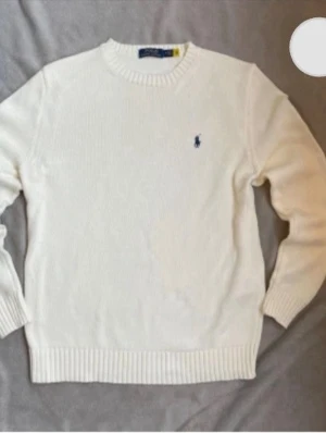 Vit stickad tröja från Ralph Lauren - Klassisk vit stickad tröja från Ralph Lauren med rund halsringning och ribbade muddar. Tröjan har den ikoniska blå logotypen broderad på bröstet och är tillverkad i mjukt material som passar perfekt till lager på lager knappt använd.