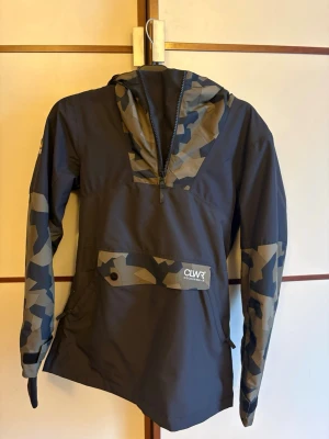 CLWR svart vindjacka med camomönster - Svart vindjacka från CLWR med camomönstrade detaljer. Dragkedjeförsedd känguruficka. Justerbar huva och half zip framtill. Ventilationsöppningar under armarna. Liftkortsficka. Lätt vadderad. Endast använd 2 gånger. Nypris: 2199kr