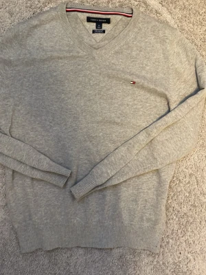 Ljusgrå stickad tröja Tommy Hilfiger - Snygg ljusgrå stickad tröja från Tommy Hilfiger med V-ringning och diskret logga broderad på bröstet. Tröjan har långa ärmar och ribbade muddar vid ärmslut och nederkant. 