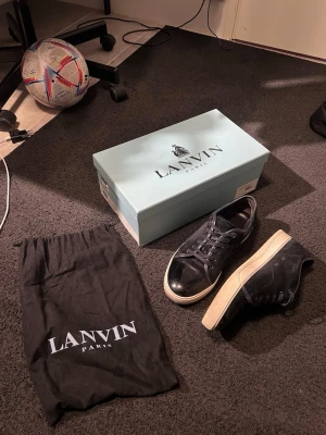 Navy Blue Lanvin sneakers med lackdetalj - Säljer ett par Navy Blue Lanvin sneakers med snygg lackad tå och vita sulor. Skorna har klassisk låg profil, svarta skosnören och är tillverkade i en mix av canvas och läder. Perfekta för dig som vill ha en clean och stilren look.