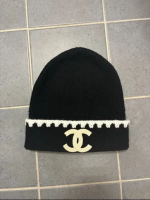 Svart mössa från Chanel med vit kant - Säljer en svart stickad mössa från Chanel med vit dekorativ kant och den klassiska Chanel-loggan framtill i vitt. Mössan har uppvikt kant och är gjord i mjukt material som håller dig varm och snygg under kalla dagar.