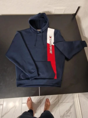 Marinblå Tommy Hilfiger hoodie - Snygg marinblå hoodie från Tommy Hilfiger med huva och dragsko. Framsidan har en vit och röd diagonal panel med logotyptryck. Klassisk känguruficka och ribbade muddar. Perfekt för en avslappnad streetwear-look.