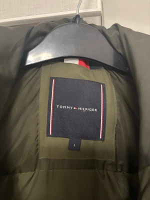 Olivgrön pufferjacka Tommy Hilfiger - Snygg olivgrön pufferjacka från Tommy Hilfiger med huva och stora fickor framtill. Jackan har diskreta logodetaljer på ärmen och bröstet samt dragkedja och knappar. Perfekt för kalla vinterdagar och riktigt trendig look.