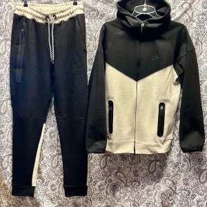 Svart och beige loungewear-set Nike - Stilrent loungewear-set från Nike i svart och beige. Setet består av en hoodie med dragkedja och två fickor samt matchande byxor med dragsko och sidofickor. Perfekt för en avslappnad och sportig look.