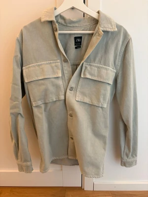 Beige overshirt från Zara i manchester - Stilren beige overshirt från Zara i mjuk manchester. Skjortan har två stora bröstfickor med lock, klassisk krage och knäppning framtill. Perfekt att bära öppen eller stängd för en avslappnad look.