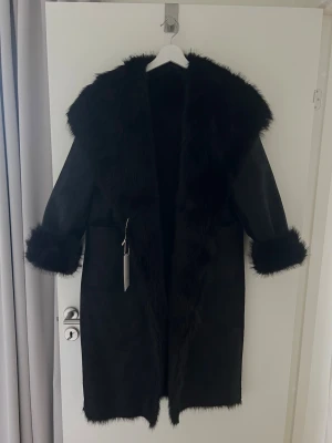 Svart vändbar Zara kappa - Snygg svart lång kappa med bred fuskpäls runt krage, ärmslut och längs öppningen. Kappan har en rak passform och är tillverkad i ett mjukt material med matt finish. Perfekt för dig som vill ha en statement piece till vinter. Det är en dubbelsidig kappa som man kan ha åt båda hållen. Ena sidan är fuskpälskappa och den andra sidan är en läderimitation med fuskpäls detaljer. Kappan är ny och helt oanvänd med prislapp kvar. 