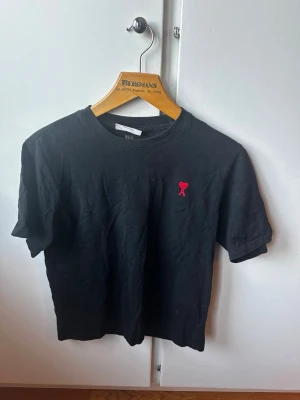 Svart t-shirt från Ami Paris - Svart t-shirt från Ami Paris med klassisk passform och korta ärmar. T-shirten har en liten röd broderad logga med hjärtdetalj på bröstet. Tillverkad i mjuk bomull och har rund halsringning. Perfekt basplagg med stilren design.