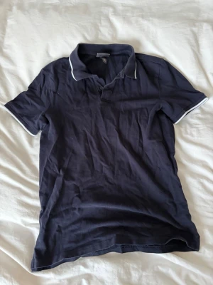 Mörkblå pikétröja - En mörkblå pikétröja med vit kant på krage och ärmslut. Tröjan har korta ärmar, klassisk krage och knappslå framtill. Tillverkad i mjukt material som känns skönt mot huden. Enkel och stilren design som funkar till det mesta.