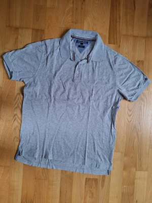 Grå pikétröja från Tommy Hilfiger - Klassisk grå piké t-shirt från Tommy Hilfiger med korta ärmar och krage. Diskreta ränder i rött och blått på insidan av kragen. Tillverkad i mjuk bomull, och otroligt skön passform. Inga defekter utöver lite veck som kan strykas bort. Nyskick, knappt använd, därav det höga priset. 