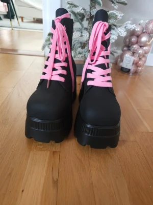Svarta boots med rosa snören, stl.41 - Säljer ett par chunky svarta boots med höga sulor och rosa skosnören. Bootsen är väldigt sköna,de har dragkedjor på insidan, rosa dödskalle- logotyp och de har dragflikar bak för enkel påtagning. Den höga platåsulan ger en markant profil.Perfekta för dig som vill sticka ut med färgglada detaljer. Köptes på Amazon, i hösten, men aldrig använd dem. Du ser det på bilderna! 