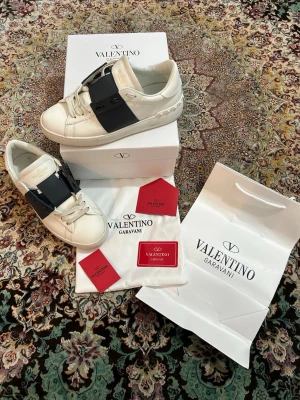 Valentino Garavani vita sneakers - Snygga vita sneakers från Valentino Garavani med bred svart rem över vristen och klassiska snören. Skorna har en chunky sula och diskret logga på plösen. Tillverkade i läder med detaljer i gummi. Perfekta för dig som vill ha en clean men exklusiv look.