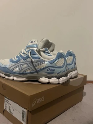 Asics nyc gel  vita och ljusblå sneakers - Snygga  oandvända sneakers från Asics i vitt och ljusblått med klassisk mesh och mockadetaljer. Skorna har en låg silhuett, vita snören och den ikoniska Asics-loggan på sidan. Perfekta för dig som gillar sportig och trendig stil.