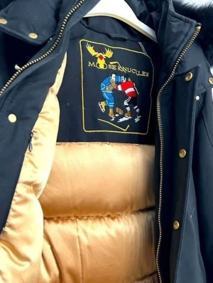 Svart dunjacka Moose Knuckles - Snygg svart dunjacka från Moose Knuckles med guldfärgade knappar och detaljer. Jackan har beige insida, avtagbar huva med svart fuskpäls och broderad logga på insidan. Perfekt för kalla vinterdagar och riktigt varm.