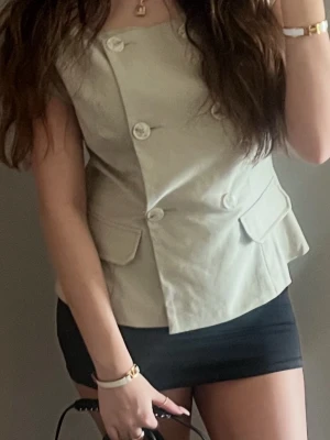 Beige asymmetrisk blus från H&M - Säljer en beige blus/trenchcoat tröja från Na-KD kollektion med Josefine HJ. Toppen har en unik skärning, två fickdetaljer och är ärmlös. Perfekt för en stilren elegant vårlook! 💖