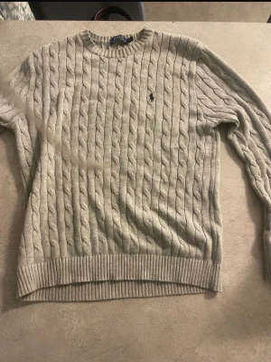 Grå kabelstickad tröja Polo Ralph Lauren - Snygg grå kabelstickad tröja från Polo Ralph Lauren med klassisk rund halsringning och svart broderad logga på bröstet. Tröjan har ribbade muddar vid ärmslut och nederkant. Perfekt för dig som gillar stilren och tidlös design.