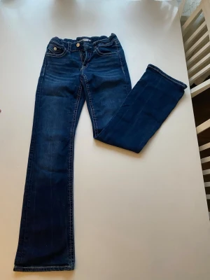 Mörkblå bootcut jeans med kontrastsömmar - Säljer ett par mörkblå bootcut jeans med vita kontrastsömmar och klassisk femficksdesign. Jeansen har knapp- och dragkedjestängning samt fickor med lock och knapp bak. Perfekta för en avslappnad och trendig look.