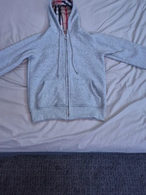 Grå zip hoodie från Burberry - Snygg grå zip hoodie från Burberry med klassiskt rutigt foder i huvan. Hoodien har dragsko i huvan, två fickor framtill och ribbade muddar. Perfekt för en chill och stilren look. Materialet känns mjukt och bekvämt.