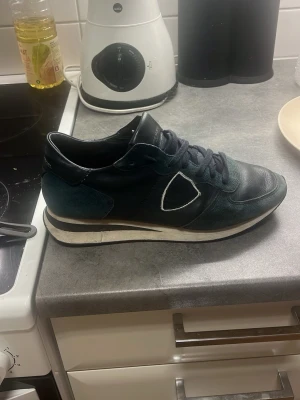 Mörkblå sneakers från Philippe Model - Snygga mörkblå sneakers från Philippe Model med vita detaljer och svart logotyp på sidan. Skorna har en klassisk låg siluett, snörning och en vit sula med svarta inslag. Materialet är en mix av läder och textil för en cool och bekväm look.