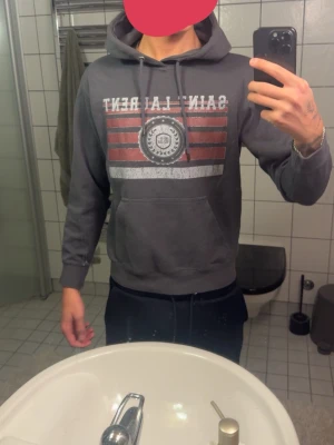 Grå hoodie Saint Laurent tryck - Säljer en grå hoodie från Saint Laurent med vitt och rött tryck framtill och stor ficka på magen. Klassisk huva med dragsko och ribbade muddar vid ärmar och midja. Perfekt för en avslappnad och cool stil.