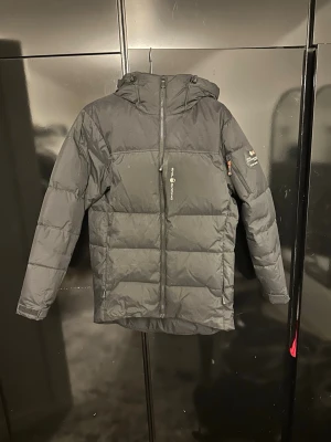 Svart Sail racing jacka - Snygg svart dunjacka från Sail racing med huva och dragkedja framtill. Jackan har en patch med logga på ena ärmen och två sidofickor med dragkedja. Perfekt för kalla vinterdagar och har en modern, puffig design.